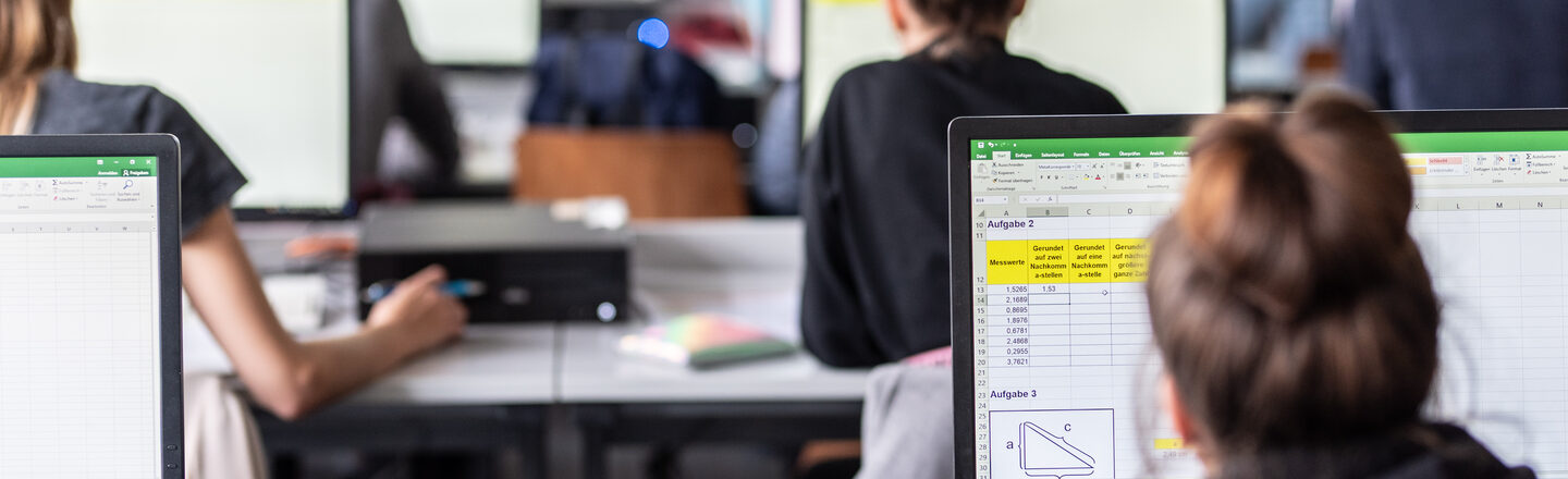 View from behind into the computer room with students, some Excel spreadsheets are visible on the monitors.