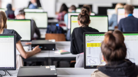 View from behind into the computer room with students, some Excel spreadsheets are visible on the monitors.