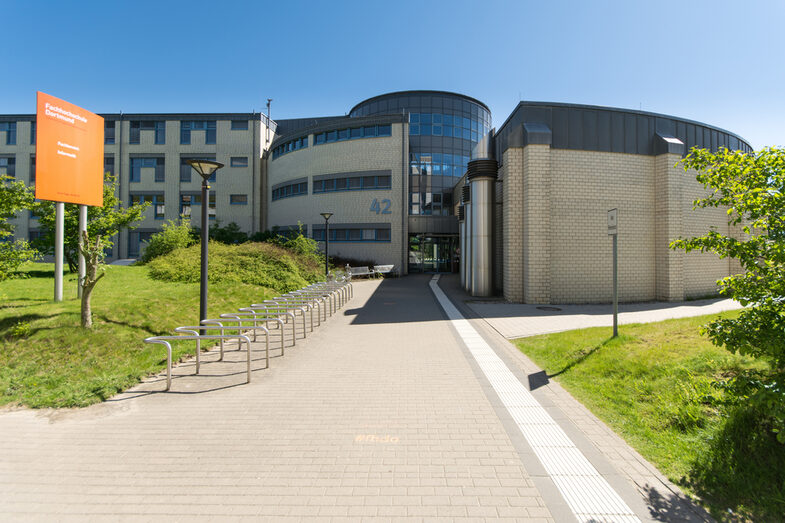 Foto des Gebäudes Emil-Figge-Straße 42 mit hinführendem Gehweg mit Blindenleitsystem. Links ein Schild mit der Aufschrift "Fachhochschule Dortmund - Fachbereich Informatik".