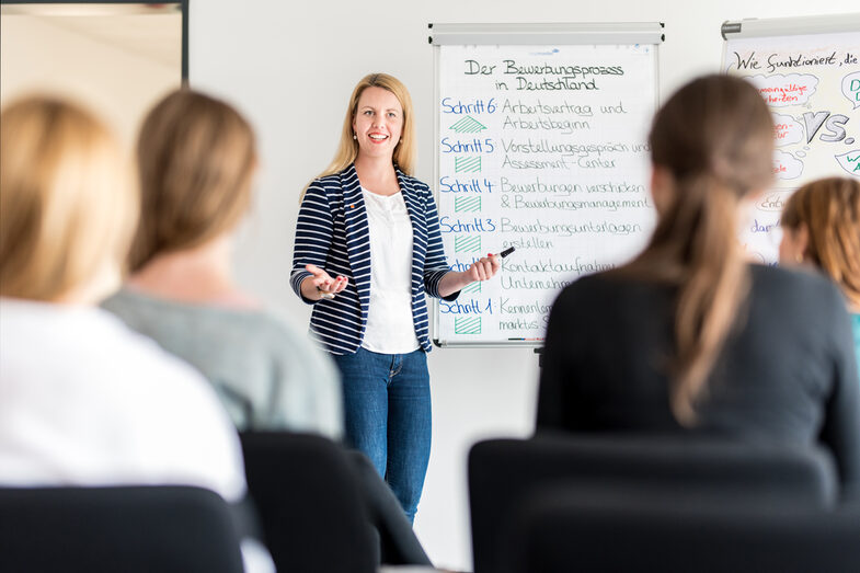 Foto einer Mitarbeiterin aus dem Career Service, die vor einem Flipchart steht, auf dem der Bewerbungsprozess in Deutschland skizziert ist. Vor ihr sitzen mehrere Studierende, die ihr zuhören.