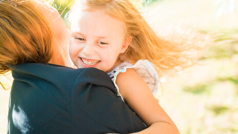 Foto einer Frau, von der man das Gesicht nicht sehen kann, mir ihrer kleinen Tochter auf dem Arm, welche freudig lacht.__Photo of a woman, whose face cannot be seen, with her little daughter in her arms, laughing joyfully.