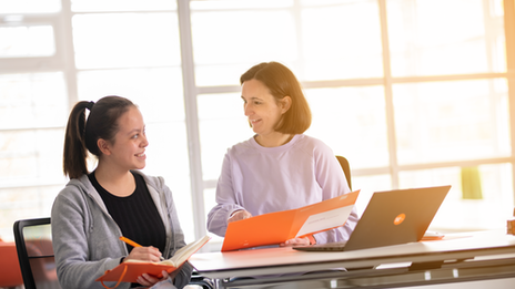 Photo of a member of the student advisory service advising a student sitting at the table next to her. She points to the folder she is holding in her hand. Both look at each other with a smile.