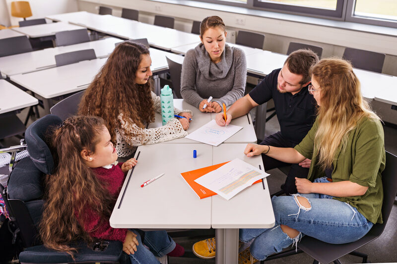 Foto einer Gruppe von Studierenden – mit und ohne Behinderungen – die an einem Tisch an einem Projekt arbeitet.