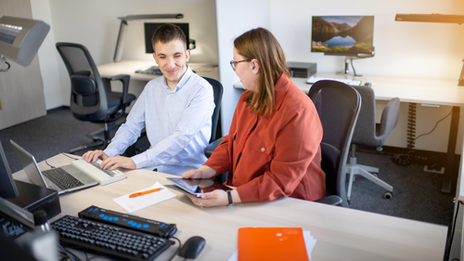 Photo of two people sitting together at an accessible workstation. The student has his hands on a Braille display, the student next to him has a tablet in both hands and is looking at the student.