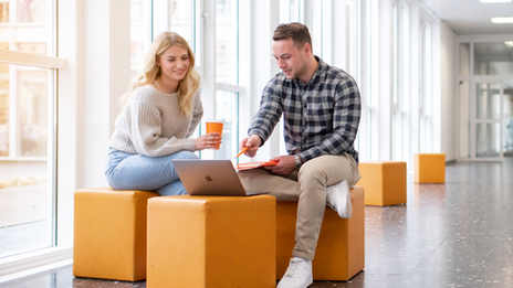 Foto von einer Studentin und einem Studenten. Sie sitzen im Flur in der FH und unterhalten sich. Der Student zeigt auf etwas am Laptop.