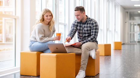 Photo of a female student and a male student. They are sitting in the hallway at the FH and talking. The student is pointing at something on his laptop.