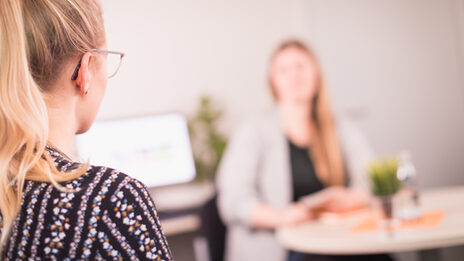 Foto einer Beratungssituation. Eine Person sitzt unscharf erkennbar an einem Tisch. Im Vordergrund ist scharf erkennbar eine Frau, ihr Gesicht ist nicht erkennbar, da seitlich fotografiert. __ <br>Counseling situation. A person is sitting at a table, out of focus. A woman is clearly recognizable in the foreground, her face is not recognizable because it is photographed from the side.