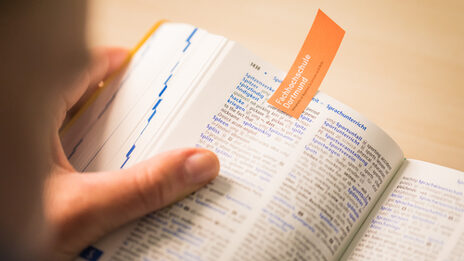 Foto eines aufgeschlagenen Wörterbuchs, das jemand in der Hand hält. Die geöffnete Seite ist mit einem orangenen Zettel mit FH-Wortmarke markiert.__Photo of an open dictionary held in someone's hand. The open page is marked with an orange slip of paper with the word mark of Fachhochschule Dortmund.