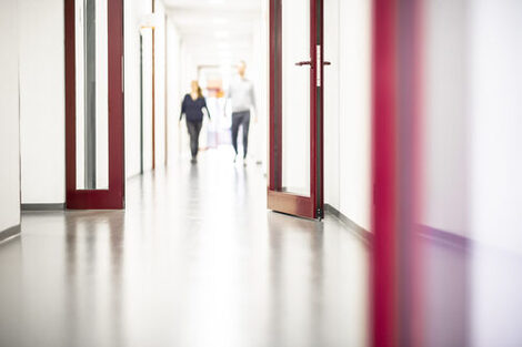 A photo of a long corridor with a red door in the foreground. Behind the door you can see two people walking along the corridor.