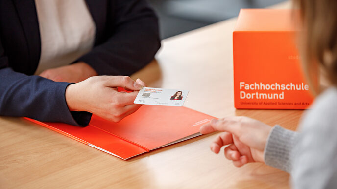 Foto von zwei Personen im Anschnitt, eine weibliche Person überreicht einer anderen Person den Studierendenausweis der FH Dortmund.<br><br>