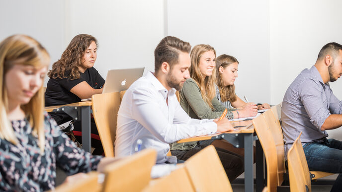 Foto in Sitzreihen eines Hörsaals, in denen sechs Studierende an Klapptischen sitzen. Der Fokus auf einer lächelnden Studentin, die anderen schauen auf die Tische.