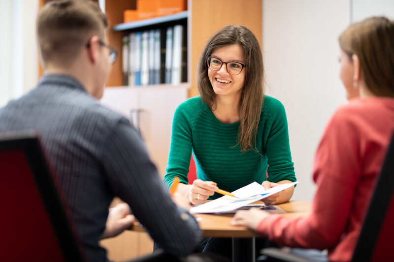 Foto einer Mitarbeiterin der Studienberatung, die mit zwei Studierenden an einem Tisch spricht. Sie hält ihnen einen Zettel hin und zeigt mit einem Stift darauf.