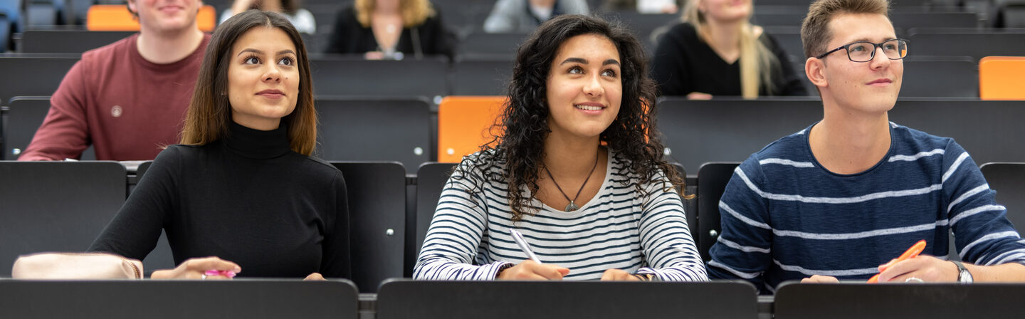 Foto von mehreren Studierenden, die in den Sitzreihen eines Hörsaals sitzen und leicht nach oben schauen.