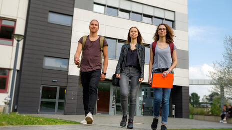 Foto von zwei Studentinnen und einem Studenten, die nebeneinander auf einem Weg gehen, im Hintergrund ein Gebäude in der Emil-Figge-Straße und weitere Studierende.__Two students walk side by side on a path, in the background a building on Emil-Figge-Straße and other students.