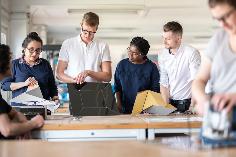 Photo of a lab member holding a metal object in his hands that is to be folded. Other students are standing around him and the workbench, watching the two of them. In the foreground, out of focus, is another student working on something.