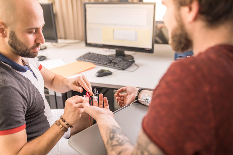 Photo of two men at a desk. One man attaches electrodes to the fingers of the other to measure his eye movements using eye tracking