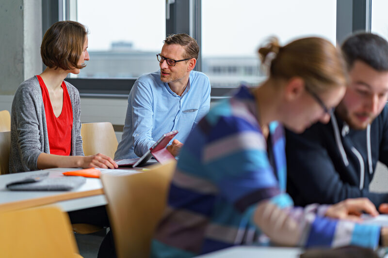 Foto von zwei Personen, die nebeneinander in einem Seminarraum sitzen und sich unterhalten. Im Vordergrund zwei weitere Personen.