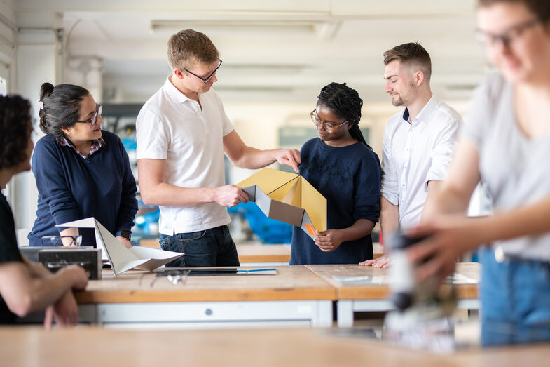 Photo of a lab member helping a student to fold a metal object. Next to them are other students watching the two. Another student in the foreground is working. __ <br>A laboratory worker helps a student to fold a metal object. There are other students standing around him and around the workbench, watching the two of them. Another student is out of focus in the foreground and is doing something.