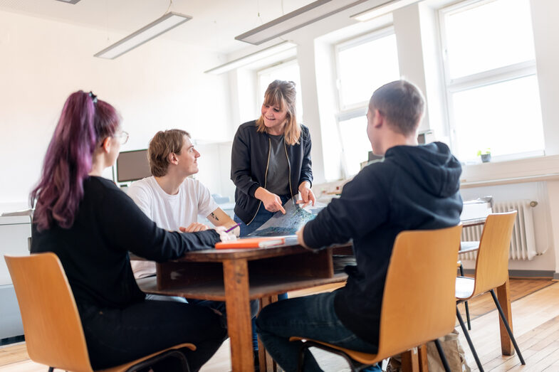 Photo of 4 people in a meeting. One person standing is showing a draft. The 3 other people sit at a table and maintain eye contact with the standing person.