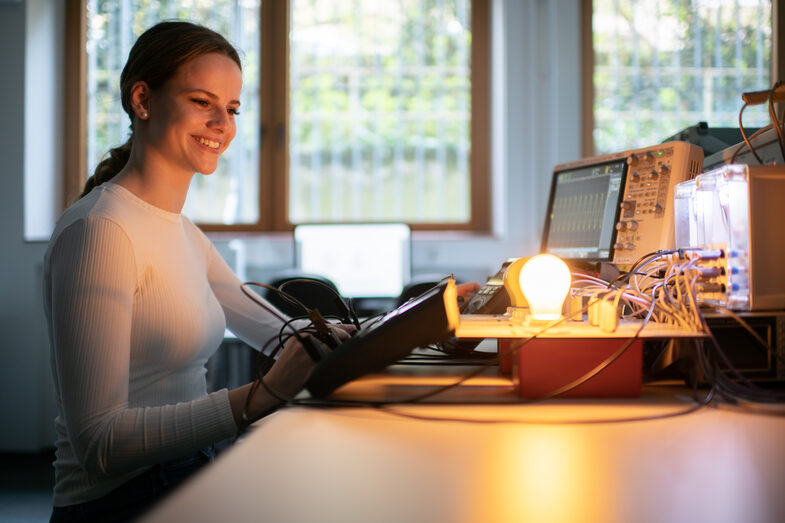 A photo of a woman sitting in front of an experimental setup. She is operating a voltmeter with her right hand. Behind the voltmeters are four lamps, one of which is lit. An oscilloscope can be seen in the background. The picture is dark because the only light comes from the illuminated lamp.