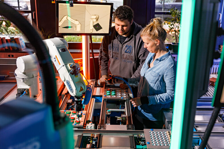 Photo of two people adjusting something on a conveyor belt with an associated robot arm.