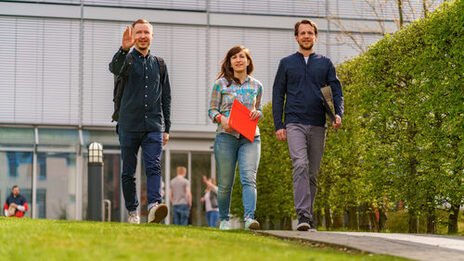 Photo of a female student and two male students walking side by side on the campus. One student waves to someone __Three students walk side by side on a path, the student on the left raises his hand in greeting.