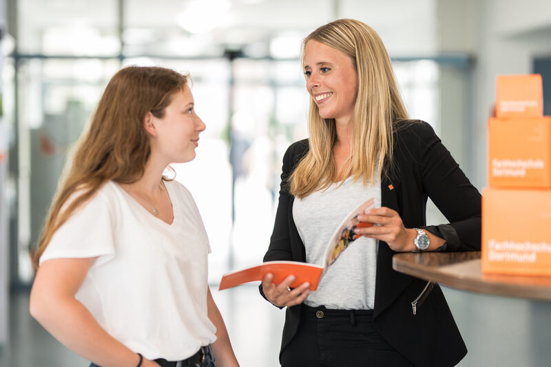 Foto von einer Schülerin und einer Mitarbeiterin der Fachhochschule Dortmund, die nebeneinander an einem Messestand stehen und ein Beratungsgespräch führen. Die Frau hält eine Broschüre in der Hand.