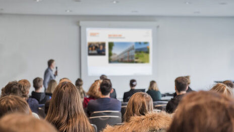 Photo of several people sitting in the rows of a lecture room, a person speaking with a microphone is out of focus at the front. __ <br>Several people sitting in the rows of a lecture room, a person speaking with a microphone is out of focus at the front.