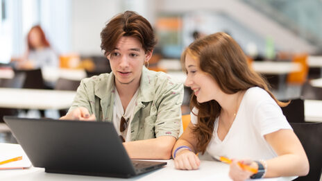 Foto von einer Schülerin und einem Schüler, die nebeneinander an einem Tisch sitzen und gemeinsam auf einen Laptop vor ihnen schauen.