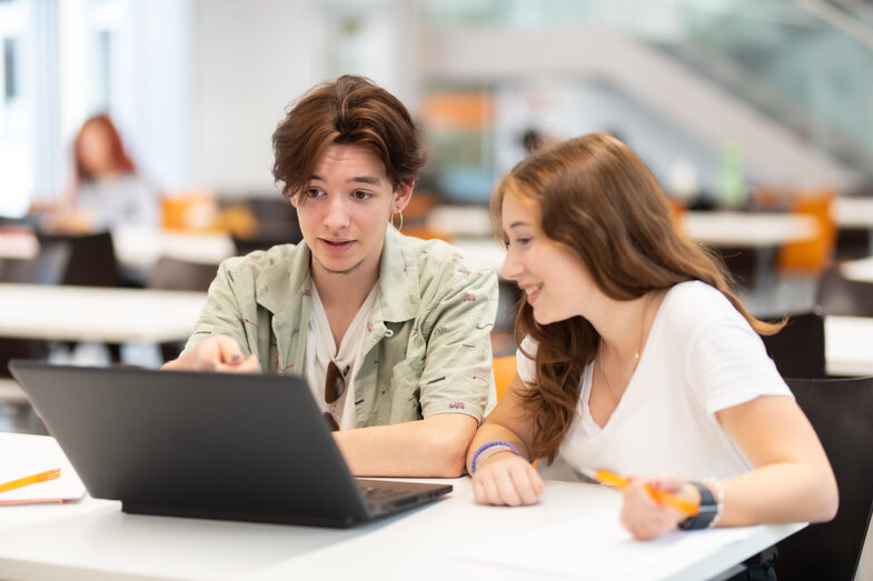 Foto von einer Schülerin und einem Schüler, die nebeneinander an einem Tisch sitzen und gemeinsam auf einen Laptop vor ihnen schauen.