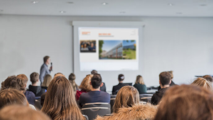 Foto von mehreren Personen in den Reihen eines Vorlesungsraumes, vorne ist unscharf erkennbar eine vortragende Person mit Mikrofon. __ <br>Several people sitting in the rows of a lecture room, a person speaking with a microphone is out of focus at the front.