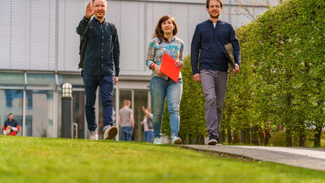 Foto von einer Studentin und zwei Studenten, die nebeneinander über den Campus gehen. Der eine Student winkt jemandem zu.__Three students walk side by side on a path, the student on the left raises his hand in greeting.