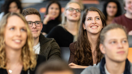 Nahaufnahme mehrerer Studierender in den Sitzreihen eines Hörsaals. Im Fokus eine Studentin und ein Student, die nebeneinander sitzen und geradeaus nach oben schauen.