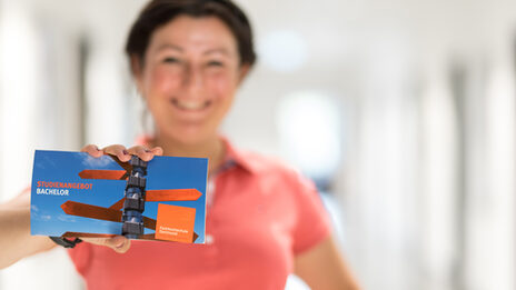 Foto einer Postkarte der Studienberatung, auf der ein Wegweiser sowie die Worte „Studienangebot Bachelor“ abgedruckt sind. Eine Frau hält die Karte in die Kamera.