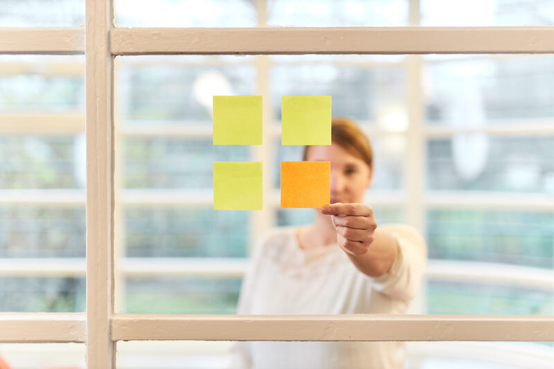 Foto einer Frau in Frontalansicht, die hinter einer Scheibe steht, an der vier quadratische Post-its kleben, die wiederum ein Quadrat ergeben. Die Frau greift an den einzigen orangefarbenen Post-it.