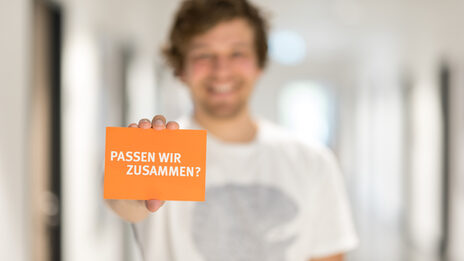 Foto eines Mannes, der eine Karte mit Schriftzug "Passen wir zusammen?" in die Kamera hält.__Man out of focus holding a card with the words "Do we fit together?" into the camera.