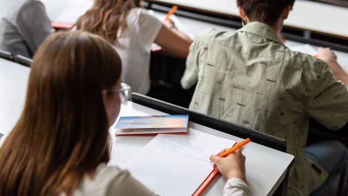 Photo of a young woman taking notes in the lecture hall. Three other people are seated in front of her. __ Young woman takes notes in the lecture hall, three other people are seated in front of her.