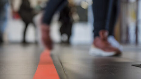 Photo of an orange line on the floor and feet of several people walking down the hall. __ <br>Orange line on the floor and feet of several people walking down the hall.