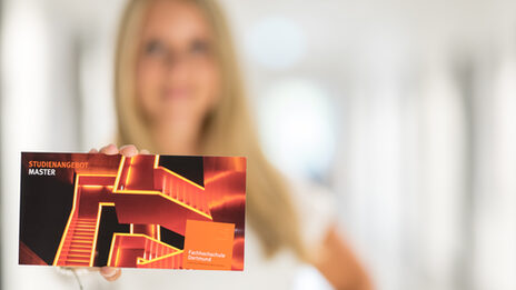 Foto einer Postkarte der Studienberatung, auf der beleuchtete Treppen sowie die Worte „Studienangebot Master“ abgedruckt sind. Eine Frau hält die Karte in die Kamera.