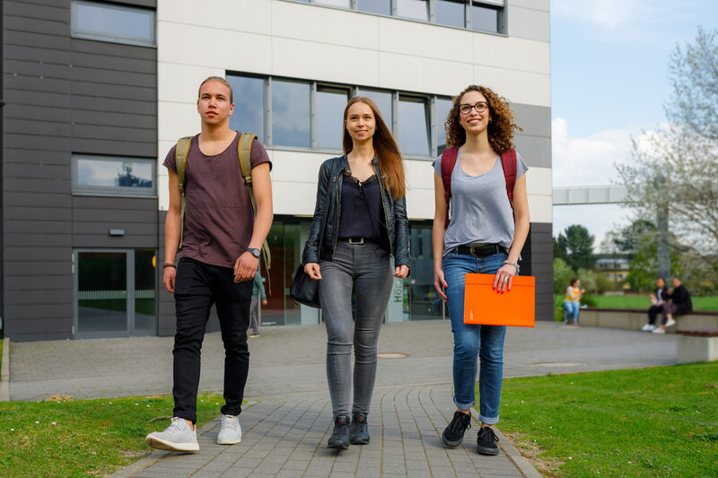 Foto von zwei Studentinnen und einem Studenten, die nebeneinander auf einem Weg gehen, im Hintergrund ein Gebäude in der Emil-Figge-Straße und weitere Studierende.__Two students walk side by side on a path, in the background a building on Emil-Figge-Straße and other students.