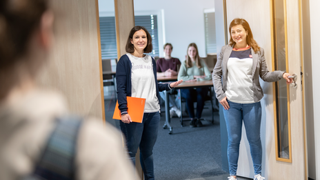 Foto von einer Studentin, die links im Bild steht, Sie ist mit dem Rücken zur Kamera. Sie schaut zur Tür ihr gegenüber, wo zwei Mitarbeiterinnen stehen und Sie herzlich begrüßen. Im Hintergrund ist ein Seminarraum mit weiteren Personen unscharf erkennbar.