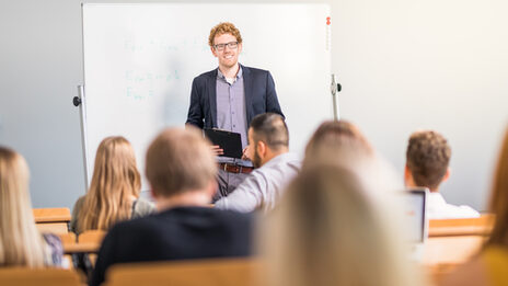 Photo of a lecturer in a lecture hall standing in front of a whiteboard, laughing. In the foreground, several seated students are blurred and seen from behind.