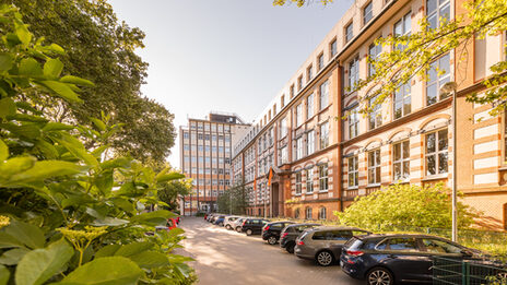 Foto des Altbaus und des Hochhauses an der Sonnenstraße mit Parkplatz davor. Links im Bild im Anschnitt ein blühender Busch.