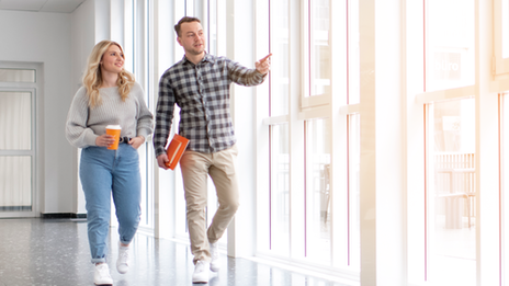 Student advisor guides Bachelor students through a corridor. The corridor is very bright and has many high windows. The student advisor points outside and explains something about the campus to the Bachelor's students.