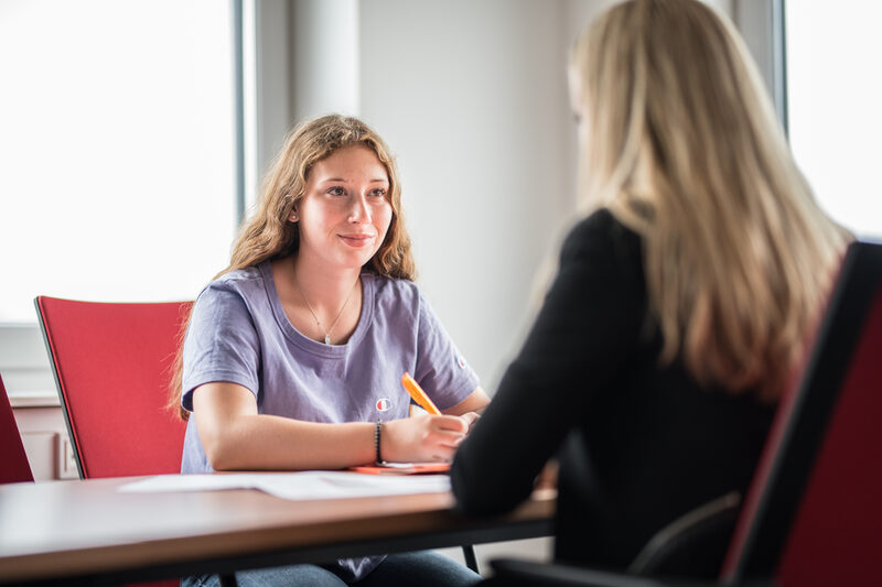 Foto einer Schülerin, die einer weiteren Frau gegenüber sitzt, welche rechts im Bild nur im Anschnitt zu erkennen ist. Sie führen ein Beratungsgespräch zur Studienberatung.