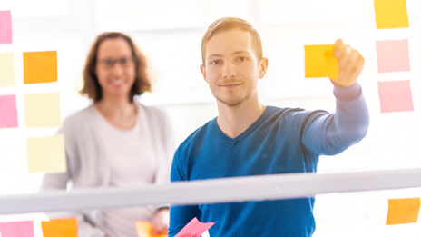 Foto von einem Studenten und einer Mitarbeiterin der Studienfinanzierung. Der Student steht im Vordergrund und klebt Post Its auf.