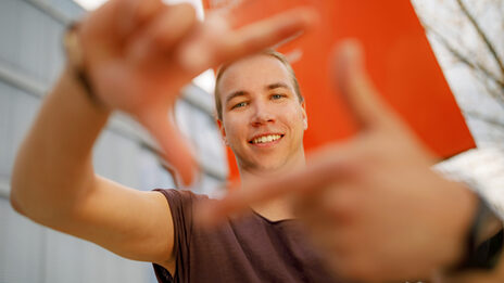 Foto eines Studenten, der mit Zeigefingern und Daumen vor sich ein Quadrat formt und dadurch guckt.  .