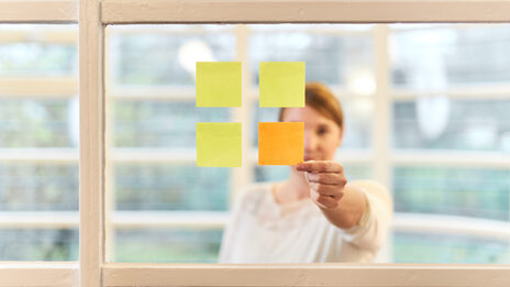 Foto einer Frau in Frontalansicht, die hinter einer Scheibe steht, an der vier quadratische Post-its kleben, die wiederum ein Quadrat ergeben. Die Frau greift an den einzigen orangefarbenen Post-it.