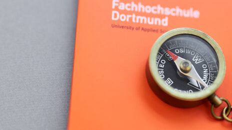 Foto eines kleinen Kompasses mit Kette daran, der mit einem Bleistift auf einer orangefarbenen Mappe der Fachhochschule Dortmund liegt.