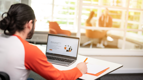 Photo of a student sitting at a laptop and taking notes. The student can only be seen from behind. In the background you can vaguely make out people in conversation.